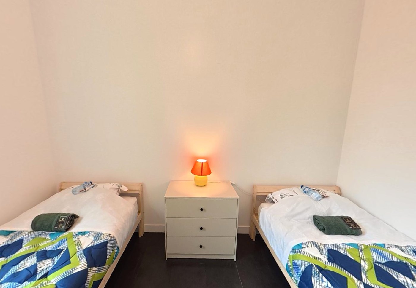 Bright bedroom with two made single beds, a chest of drawers and a bedside lamp